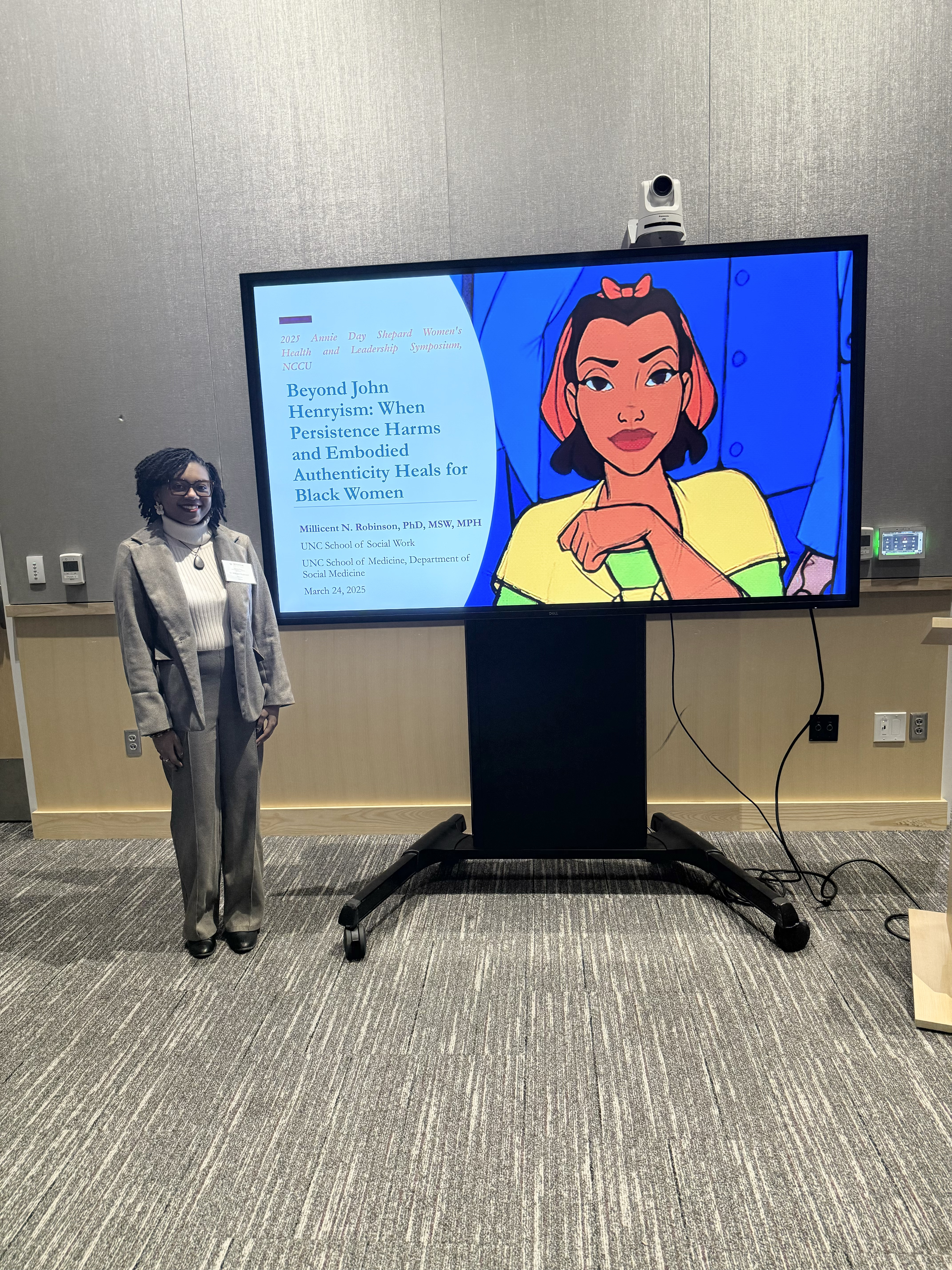 A Black woman in a gray suit stands next to a digital screen displaying a presentation titled 'Beyond John Henryism: When Persistence Harms and Embodied Authenticity Heals for Black Women.' The screen also features an illustrated character.