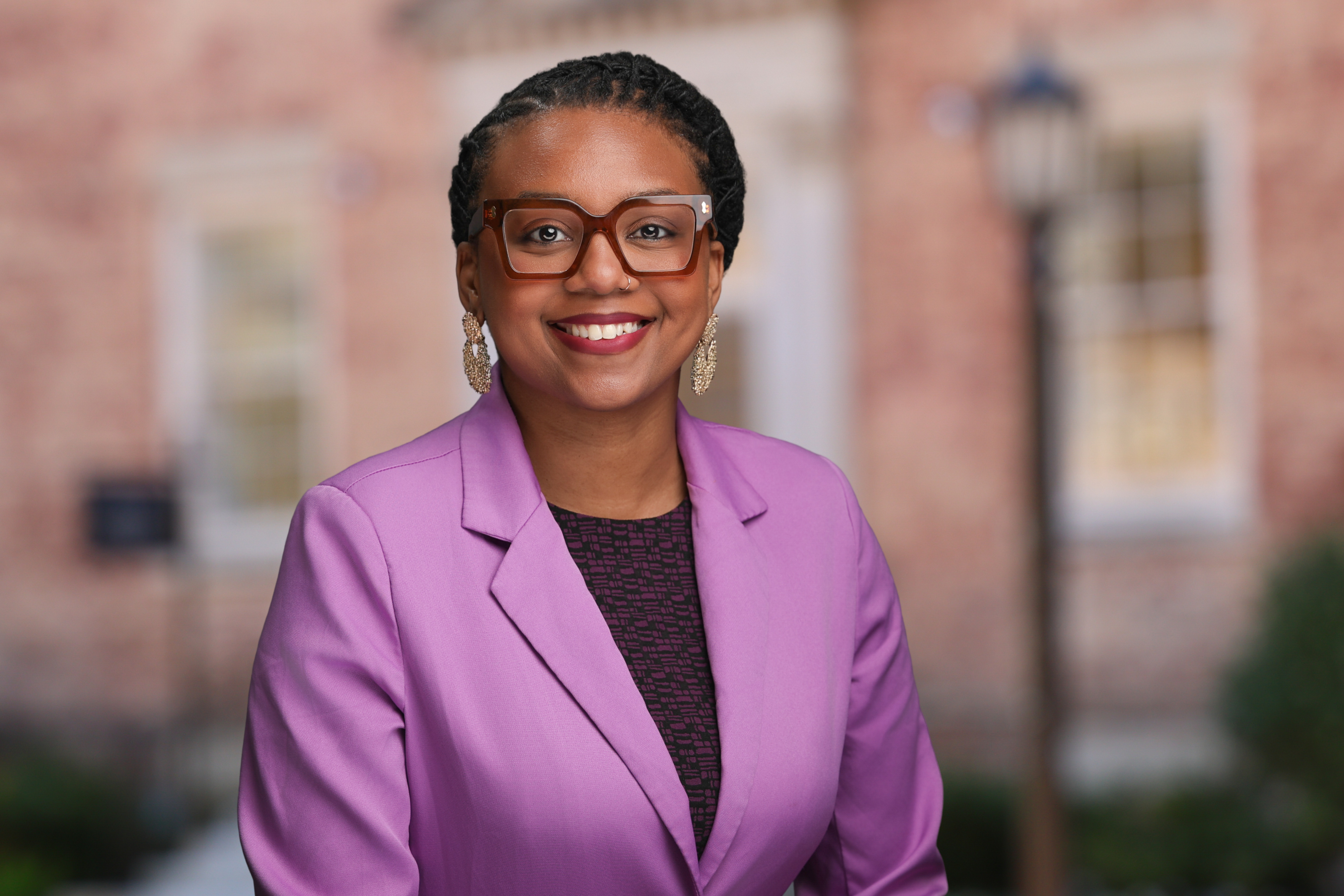 Black woman smiling, wearing glasses, a purple dress and blazer in a professional headshot.