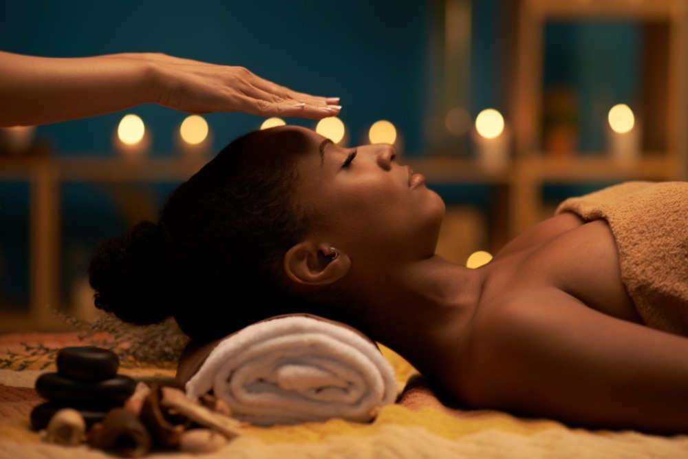 Picture of Black woman lying down with hands hovering over her face with lights in the background to illustrate reiki.
